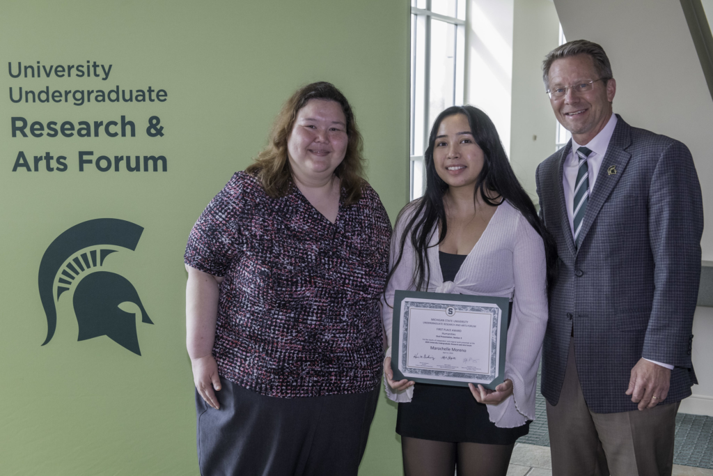 A group of three people stands in front of a green banner with the text "University Undergraduate Research & Arts Forum" featuring a spartan helmet logo. From left to right: a woman with shoulder-length hair, wearing a printed blouse and black bottoms; a woman holding a certificate, with long black hair, wearing a light cardigan over a black dress; and a man in a gray patterned suit with a spartan helmet logo pin on his lapel, all smiling at the camera.