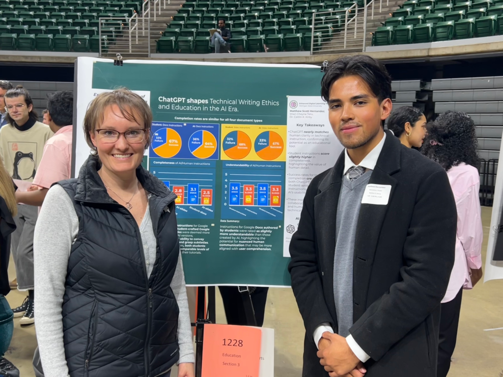 Two individuals stand in front of a research poster presentation. On the left is a woman with short hair, wearing glasses, a gray long-sleeve shirt, and a black vest. On the right is a young man with styled dark hair, wearing a black coat over a white shirt and tie, with a name tag. The poster is about "ChatGPT shapes Technical Writing Ethics and Education in the AI Era." They are both smiling at the camera, with an audience and other presenters in the background in an indoor arena setting.